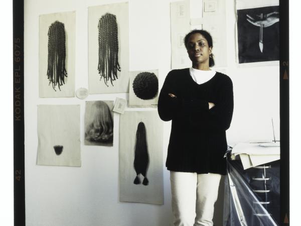 Photo of Lorna Simpson in her studio in Brooklyn, New York. She crosses her arms and stares at the camera. Behind her are her prints of different African American hairstyles.