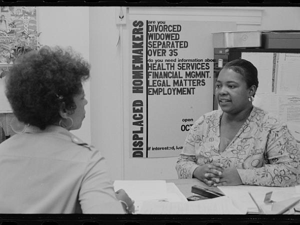 Two women seated at a table, the woman in front of the other woman has her hands folded and a seams to be mid conversation. There is a poster in the background that reads "DISPLACED HOMEMAKERS are you DIVORCED WIDOWED SEPARATED OVER 35 do you need information about HEALTH SERVICES FINANCIAL MGMNT. LEGAL MATTERS EMPLOYMENT 