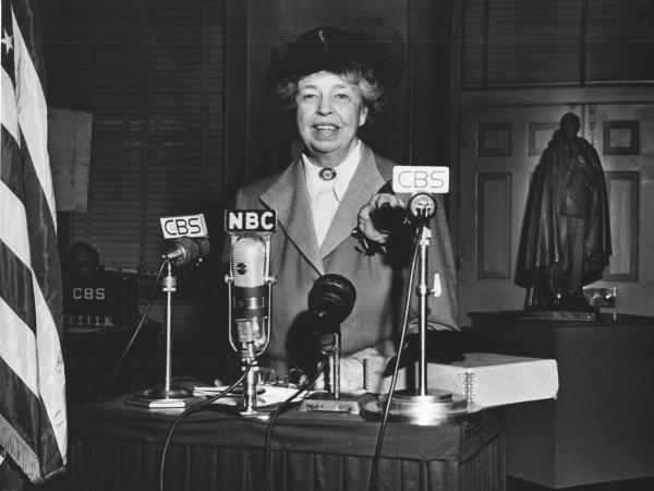 Eleanor Roosevelt standing in front of four microphones labeled as CBS and NBC. 