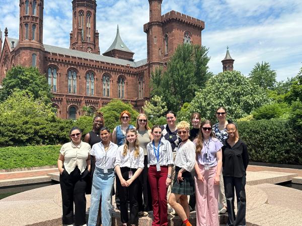 A group of thirteen people stand together in front of green shrubs and the Smithsonian Castle. 