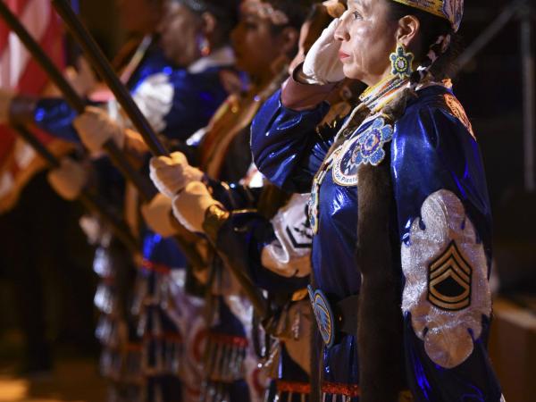 Mitchelene BigMan and a line of other Native American Women Warriors holds a flag. They all wear ceremonial dresses.