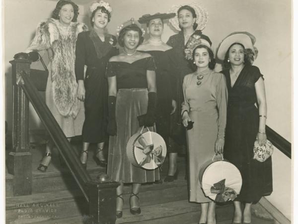 A black-and-white photograph of seven women standing on a flight of stairs. Mae Reeves stands to the farthest right on the lowest step and wears a white hat and floral-patterned handbag.