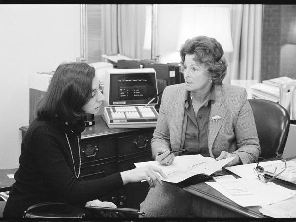 A woman in a suit behind a desk with a pen and documents in hand. Another woman in a turtleneck and pearls sits in front of her and is pointing to the document. 