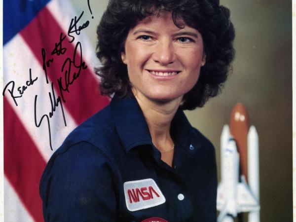 Autographed posed photo of Sally Ride. She wears a polo with a NASA patch and a Challenger patch. She stands in front of an American flag.