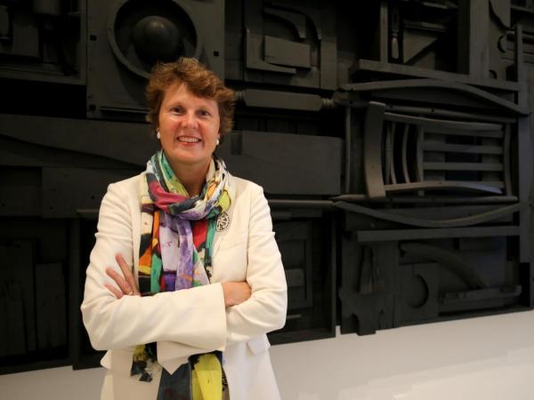 Stephanie Stebich stands in front of a wall-mounted artwork with wood objects painted black