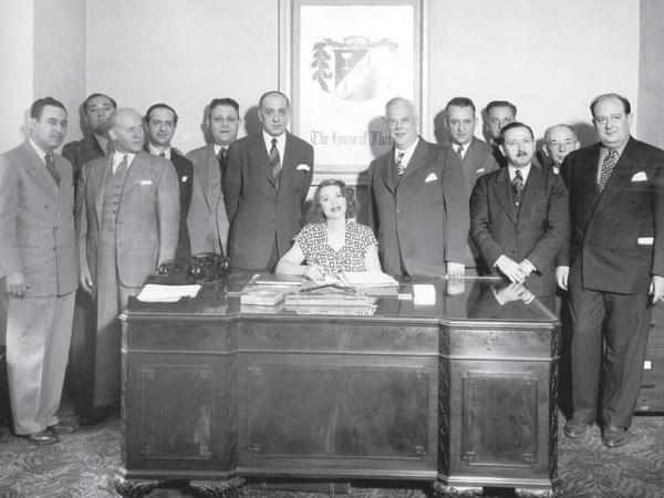 Tillie Lewis sits at an office desk. She is surrounded by men in suits.