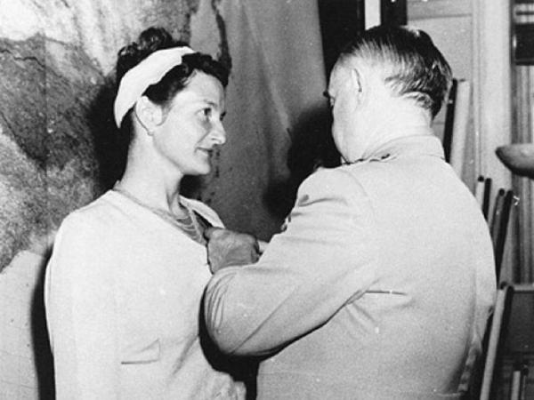 Virigina Hall looks at General Donovan as he pins the Distinguished Service Cross to her top. She wears a light colored suit with a skirt. Her hair is done in an updo with a wide light-colored hairband.