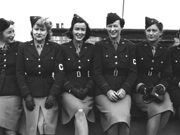 Six women sitting closely together posing for the camera. They are wearing uniforms with light skirts and dark jackets and an armband with the letter C on it.  