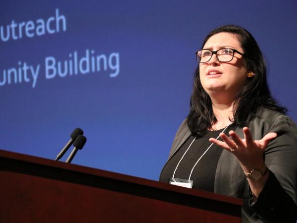 Kelly Doyle stands at a podium. She holds up her hand, gesturing as she speaks. She wears a conference badge. Behind her is a screen that says &quot;Outreach / Community Building.&quot;