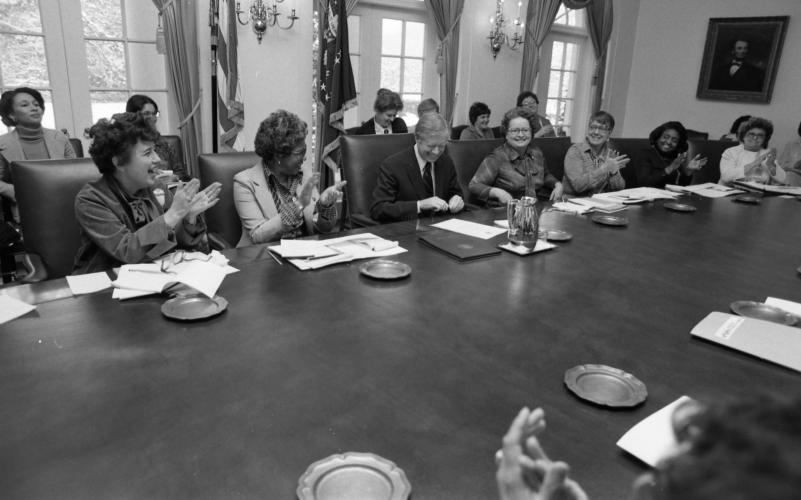 Candid black and white photo of President Carter at a long table with papers in front of him. He is smiling and closing a pen cap. A group of women is seated at the table beside him and standing behind him clapping.     