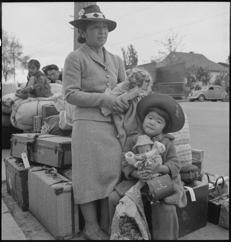 Black and white photograph of a Japanese American woman with a young girl holding a doll in front of a pile of luggage, with another Japanese American family in the background. 