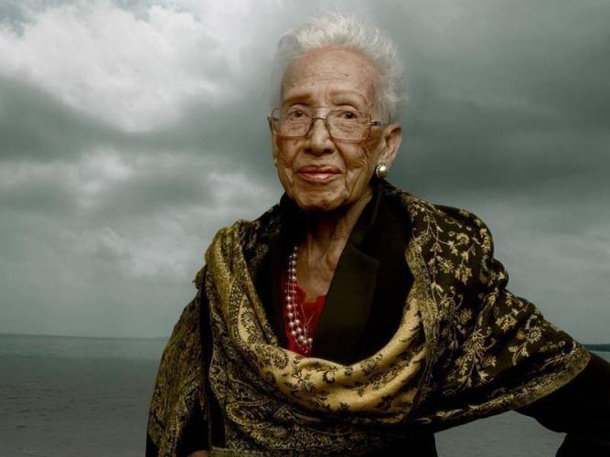 Katherine Johnson looks at the camera. She stands with one hand on her hip. She wears a gold floral scarf, a black blazer, and pearls. Gathering clouds swell behind her.