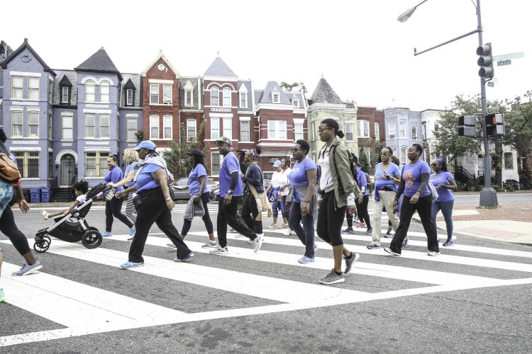 A group of people wearing similar t-shirts in a crosswalk of a street with red and blue rowhouses.