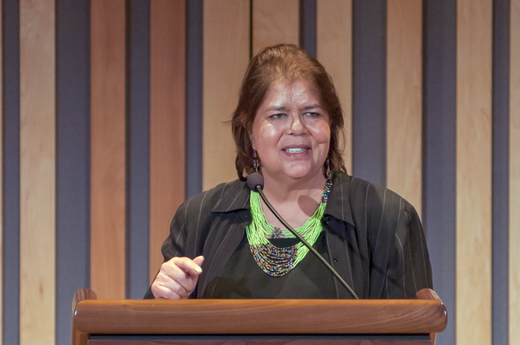 Wilma Mankiller speaking at a podium while wearing a green beaded necklace 