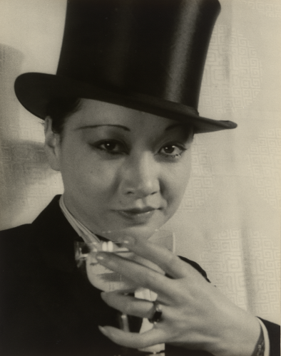 Black and white photo of Anna May Wong wearing a suit and top hat. She arches an eyebrow and holds a glass to the camera in a toast.