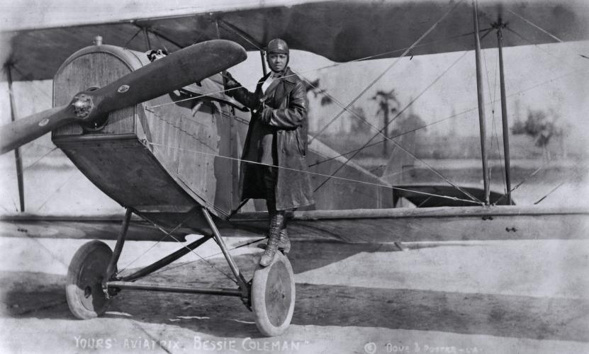 Bessie Coleman, dressed in a long aviator’s coat, hat, and boots, grips a plane’s frame while standing on its front left tire