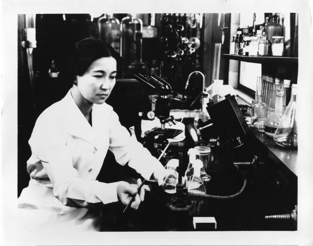 Ruby Hirose sits at a lab station. She wears a white lab coat and holds a pipette.