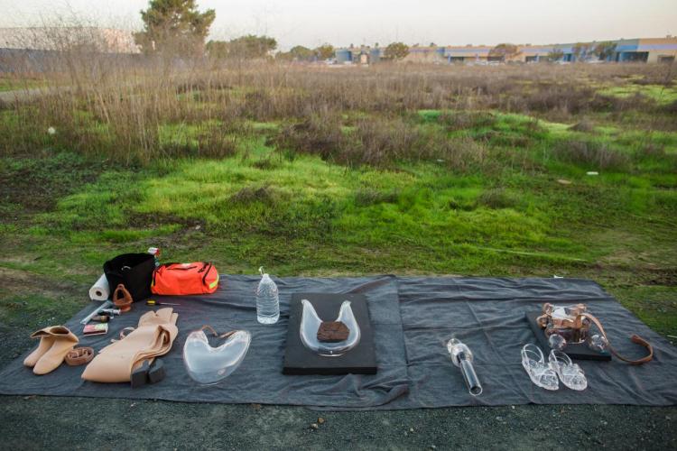 Objects belonging to the artist arrange over top of a black tarp. The tarp is outdoors in a grass covered lot. 