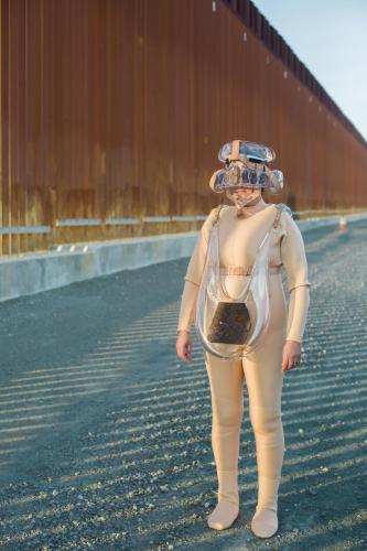 Artist wearing beige costume and standing outdoors in a vacant lot. 