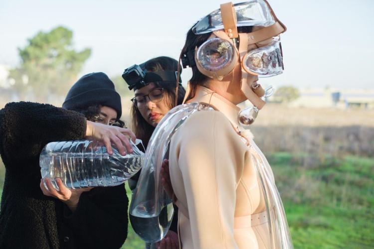 The artist in her beige costume standing with two other people. The two people are filling a jug with water that is being worn by the artist like a backpack.