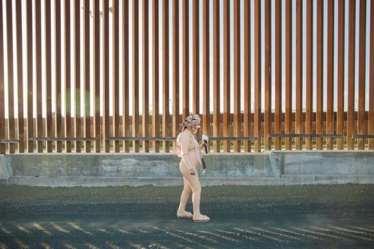 The artist standing in her costume in front of the border wall. 