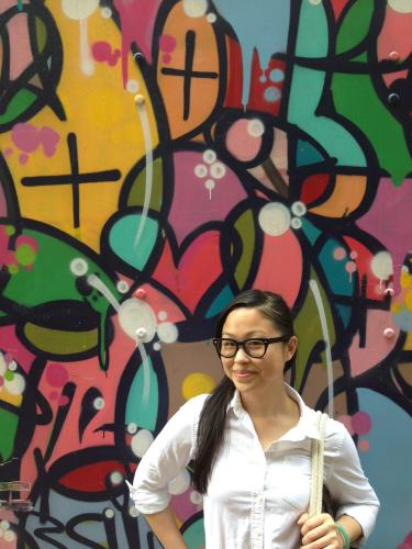 A young Vietnamese woman with dark hair and glasses wearing a white shirt stands in from of colorful wall mural.