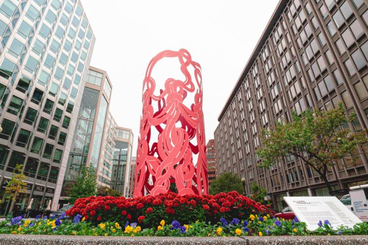 Photo of Marker, a tall cylindrical metal sculpture depicting pink intersecting threads. It stands above a bed of colorful flowers.