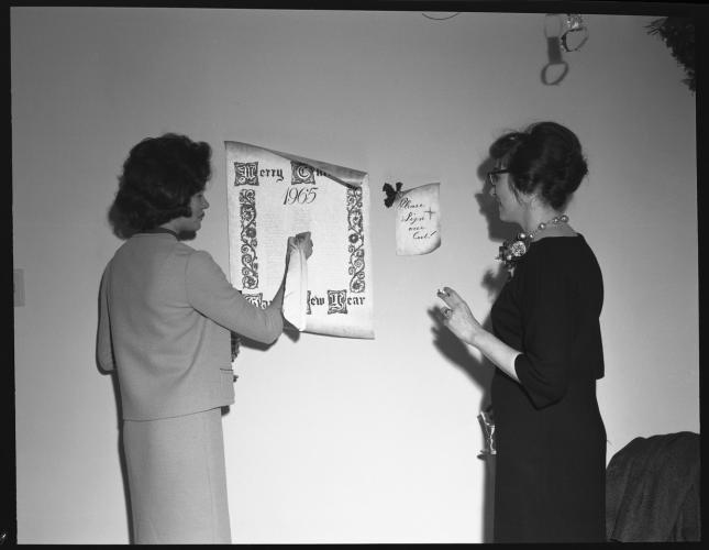 Carolyn Gast stands beside a colleague to the right. Her colleague is holding a feather quill and writing on a page stuck to the wall with decorative lettering around the border and the words “Merry Christmas 1965” at the top. 