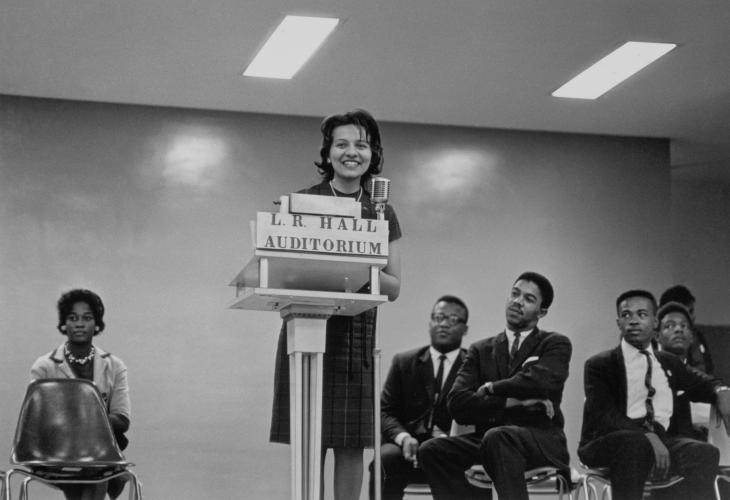 A black-and-white image of Diane Nash at the podium with men and a woman sitting on either side of her in chairs. Nash stands in the center of the image behind the podium labeled [L.R. HALL/ AUDITORIUM] speaking into the microphone.  
