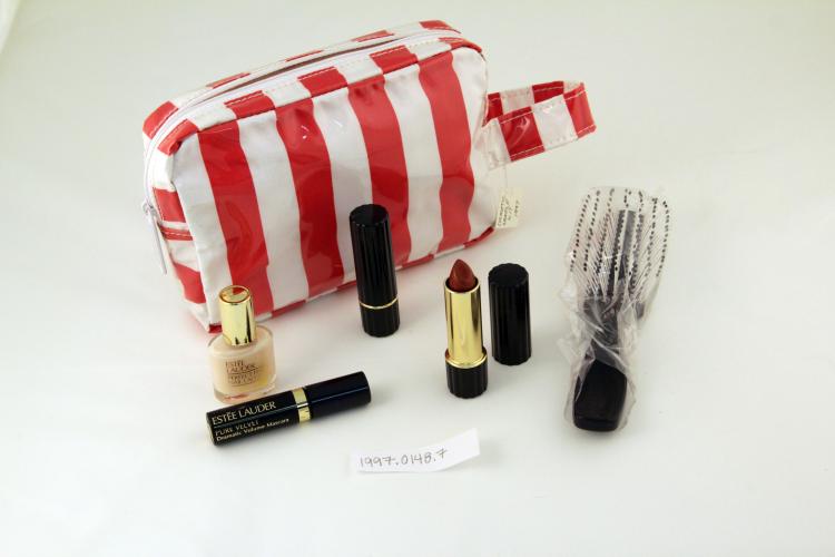 A white-and-red striped cosmetic bag surrounded by Estée Lauder products, including tubes of lipstick and a hairbrush.    