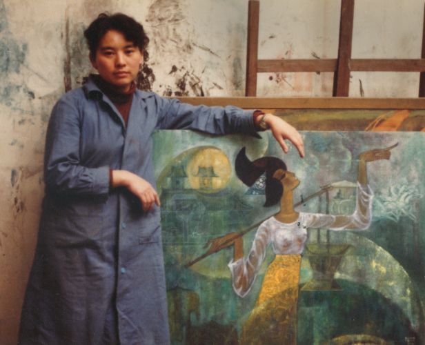 Photo of Hung Liu wearing an artist&#039;s smock and posing with a painting of a brown-skinned woman pointing off into the distance
