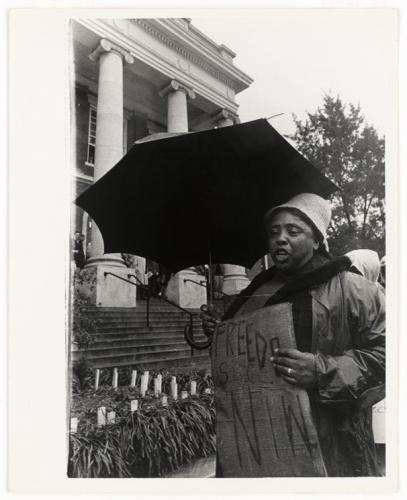 A black-and-white photograph of Fannie Lou Hamer carrying a sign and holding an umbrella over her head.