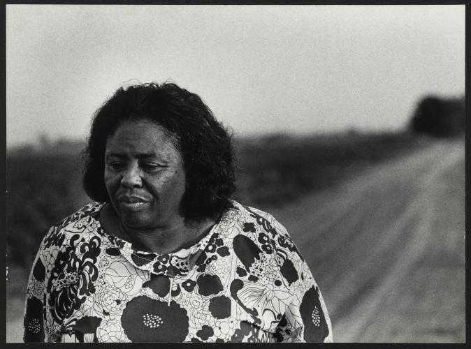 A gelatin silver print of Fannie Lou Hamer wearing a floral print top. She is standing on a dirt road and is looking off camera in the bottom left corner of the frame.  
