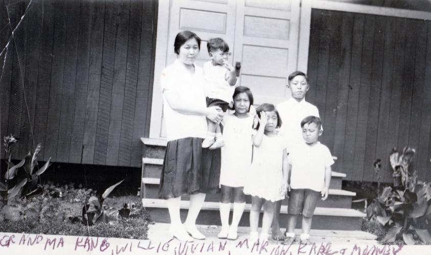 Black and white photograph of Sung Hark (Lee) Kang standing on steps in front of a house holding a young boy on her hip. Her other four children, two girls and two boys, stand to her left.    