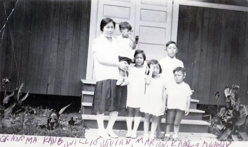Black and white photograph of Sung Hark (Lee) Kang standing on steps in front of a house holding a young boy on her hip. Her other four children, two girls and two boys, stand to her left.
