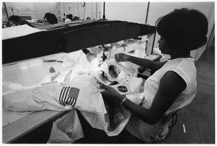 Black and white photograph of African American woman sitting at a long-arm sewing machine working on the iconic white outer layer of an Apollo spacesuit.
