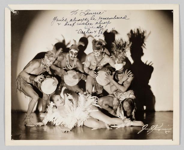  Princess Orelia lies on the floor in a feathered dress, with four men in costumes with conga drums standing behind her