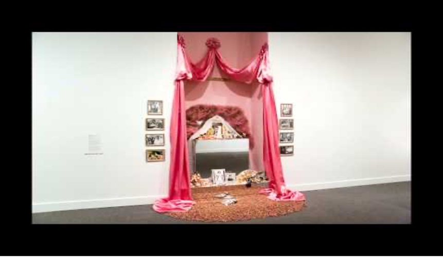 Color photograph of installation art in the form of an ofrenda.  An altar formed by coral pink drapery surrounds a makeup vanity table with a white lace and pink fabric crown. The altar is flanked by four movie stills of Dolores del Rio on each side in a vertical configuration. The vanity table holds black and white photographs, candles, souvenirs, images of the Virgin of Guadalupe and examples of Mexican folk art. Miniature, dried pink rosebuds spill out of the ofrenda onto the floor.