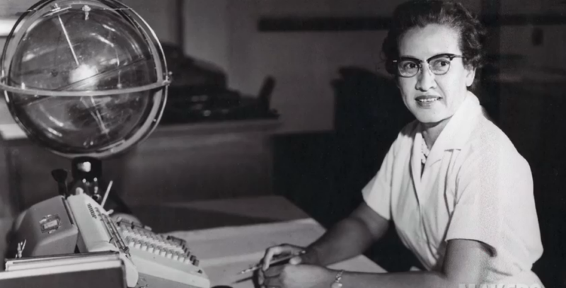 Katherine Johnson looks at the camera. She sits at a desk with papers for calculations and a large globe.