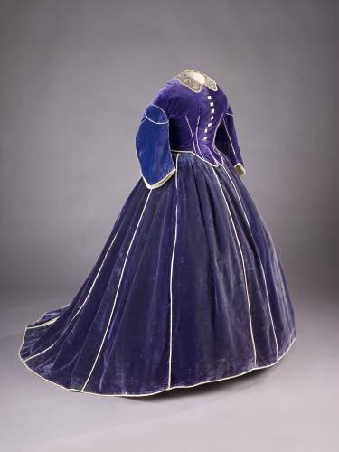 Half-profile photograph of a long-sleeve, floor-length purple velvet gown with a white lace collar on a dressmaker’s mannequin.