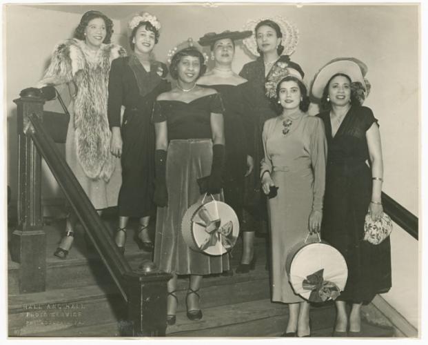 A black-and-white photograph of seven women standing on a flight of stairs. Mae Reeves stands to the farthest right on the lowest step and wears a white hat and floral-patterned handbag.