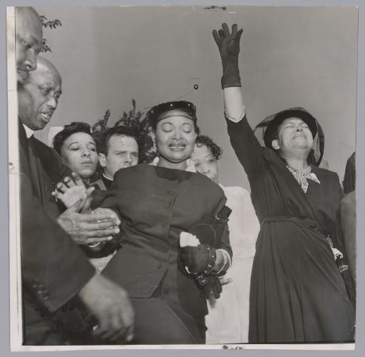 Black-and-white image of Mamie Till-Mobley standing in front of 5 people. An unnamed woman to her left has her left hand raised and is crying.  