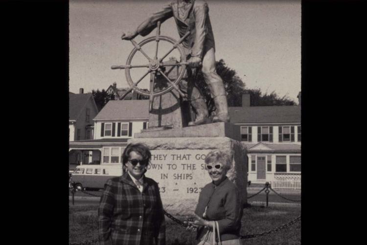 Patricia Morse and Marian Pettibone smiling in front of a tall stone statue of a sailor holding onto a ship steering wheel. 