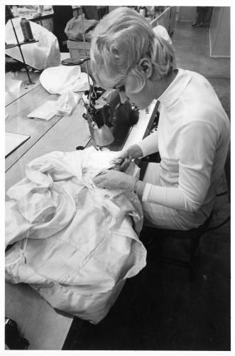 Black and white photograph of the profile of a seamstress with blonde hair and white clothing using scissors to trim threads on a white fabric component of an Apollo spacesuit.