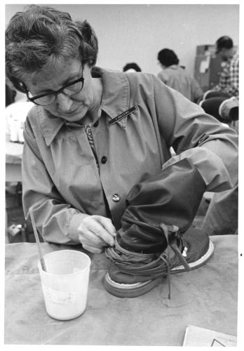 Photograph of an older woman wearing glasses and a collared smock adjusts flexible material connecting the leg and heel of a boot. A brush sits in a container of adhesive on the table in front of the woman; other workers are visible in background.
