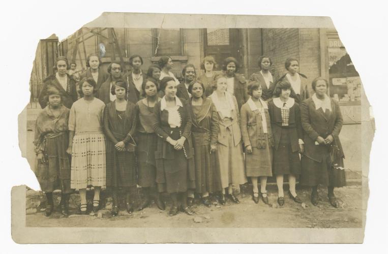 This black and white photograph that depicts twenty-one African American women and one small child. The women stand in two rows on the steps of a building.