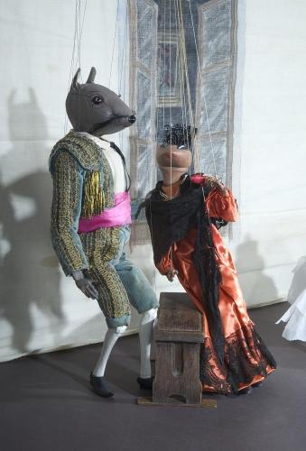 Marionette puppets of a rat and a cockroach, both dressed in fancy human clothes, posed on a stage set with a window and seat