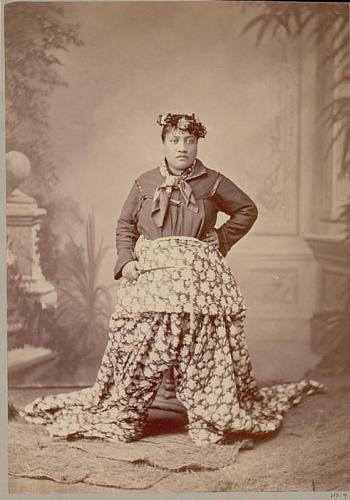 Woman wearing a floral crown, neck scarf, and long floral-printed skirt. She stands with a hand on her hip and holds a riding crop.