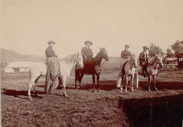 Four people on horseback in a field. There are two women wearing long patterned skirts, and two men.