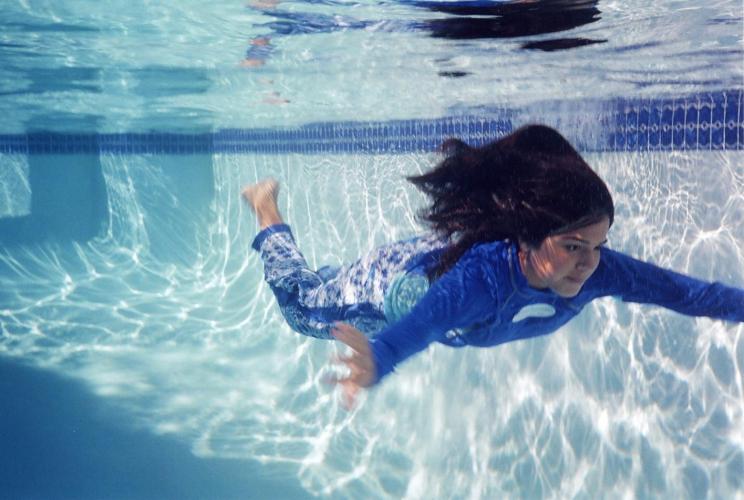 A woman swims underwater in a pool while wearing a blue and white shirt, pants, and swimhood.